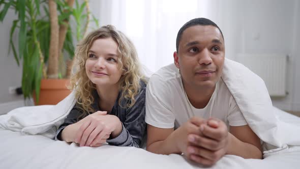 Shy Millennial Interracial Couple Looking at Each Other Smiling Lying in Bed Under Blanket alt