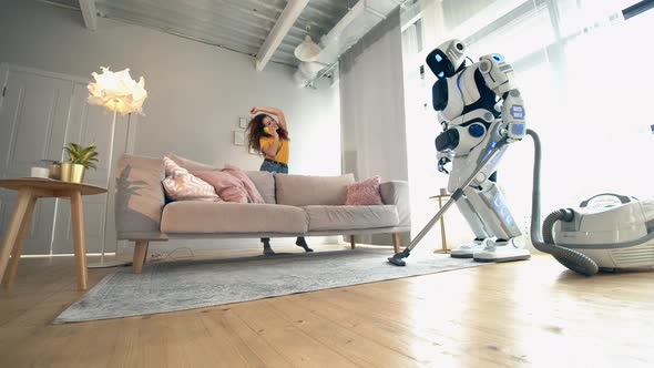 Young Woman Is Talking on the Phone While the Droid Is Hoovering. Robot, Human-like Cyborg Concept alt