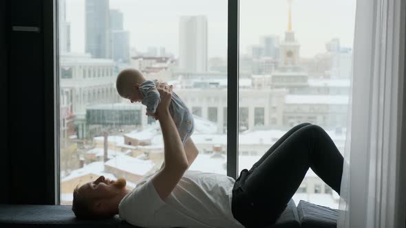 father raises the baby above him while lying on the windowsill by the window