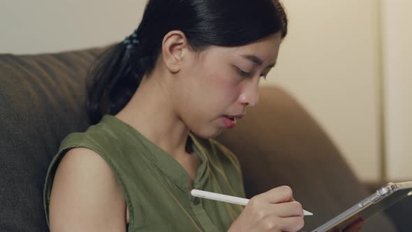 Asian female reading news and trading stock on her digital tablet while sitting on the sofa.