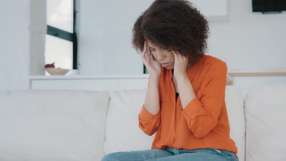 African American Girl with Curly Hair Woman Sitting at Home on Couch Touching Massages Temples Feels alt