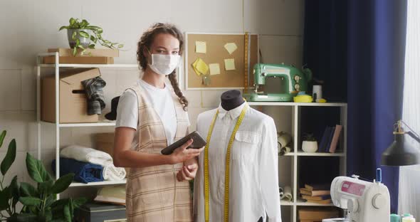 Young Woman Dressmaker Wearing Medical Mask Standing Near Mannequin in Workshop Holding Gadget alt