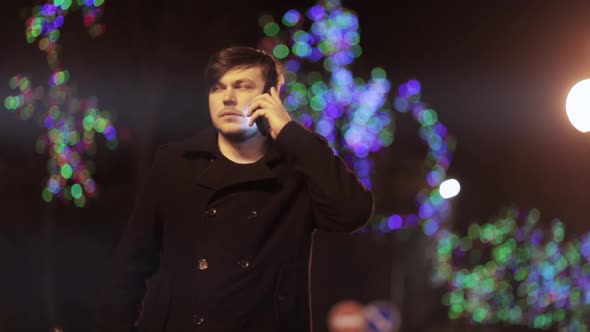 A man in a black coat talking on a smartphone while walking in the city at night