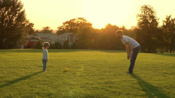 Little Child Girl and Young Father Play to Football on Green Lawn in Summer Park at Sunset alt