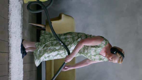 Vertical View of Cheerful Mature Housewife Using Vacuum Cleaner and Listening To Music in Headphones alt