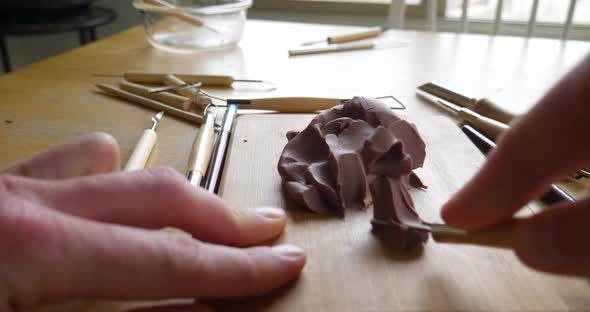 The hands of a sculptor using tools and brushes to cave and shape brown modeling clay for a sculptin alt