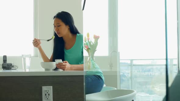 Woman using mobile phone while having breakfast in kitchen alt