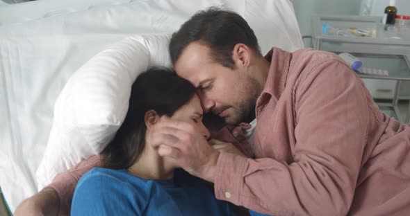 Husband Hugging and Comforting Crying Sick Woman Lying Together in Hospital Bed alt