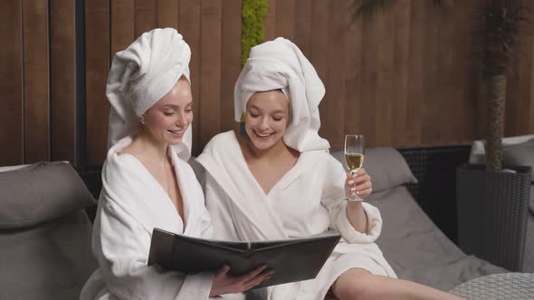 Girlfriends in Spa Salon in Towel and Bathrobe Looking at Prices of Procedures Drinking Champagne