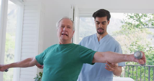 Young male physiotherapist helping retired senior man exercising with dumbbells at nursing home alt