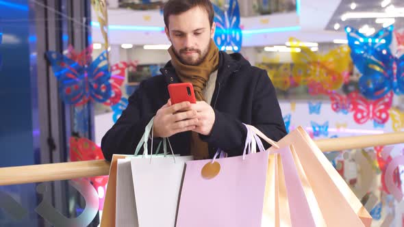 Joyful Young Man Is Chatting Online While Standing in Shopping Mall with Lots of Gifts alt