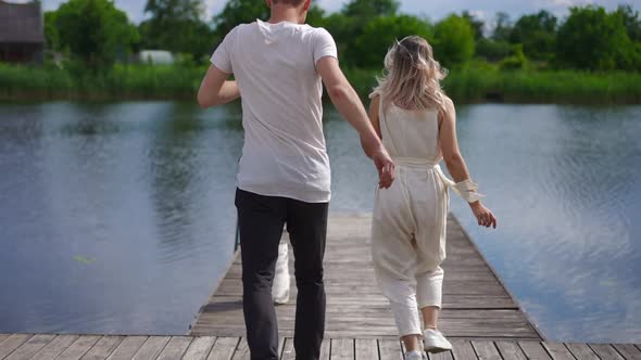 Tracking Shot of Excited Young Couple Running in Slow Motion to River on Wooden Pier Smiling alt