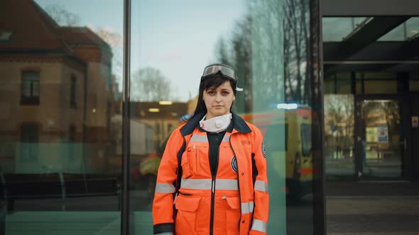 Pretty Female Paramedic Stands Near Modern Hospital and Looks at Camera alt