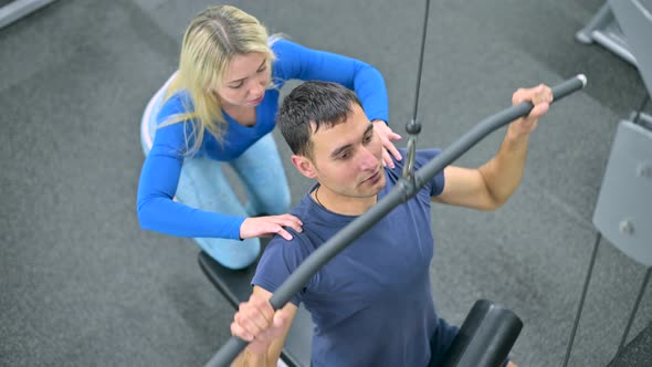 Slim guy performs an exercise on machine under the supervision of coach alt