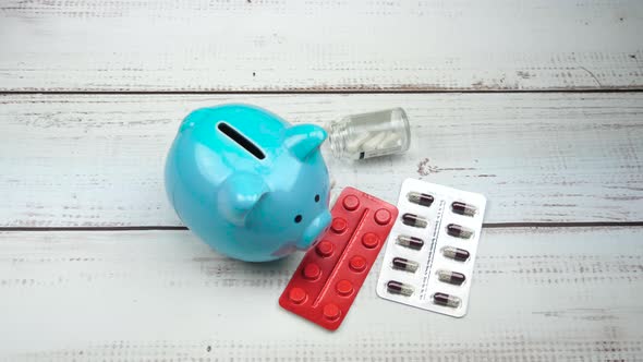 Hand Puts a Coin in the Piggy Bank. Pills, Medications Are on the Table. The Concept of Savings alt