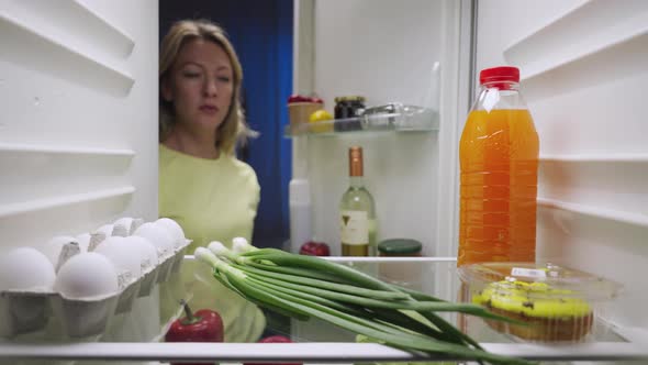 Sleepy Young Woman Opens the Refrigerator Door at Night Looks Inside and Picks Out a Bottle of White alt