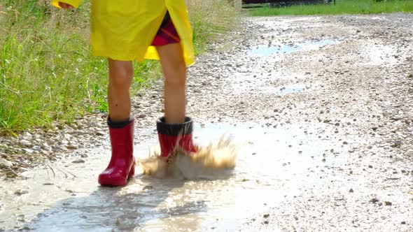 A girl in red rubber boots and a yellow raincoat runs through puddles after a rain in the village. S alt