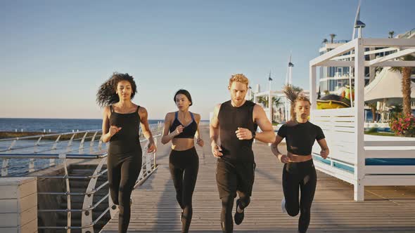Muscular Man Couch Running with Three Diverse Sporty Ladies on Pier Warming Up Before Strength alt
