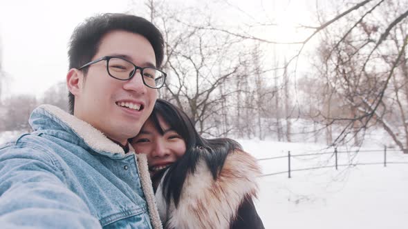Happy Young Couple Hugging and Taking Selfie in the Park on Snowy Winter Day alt
