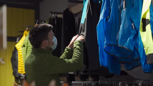 Masked Man in Sports and Outdoor Equipment Store Selects Jacket During Covid 19 Quarantine and Keeps alt
