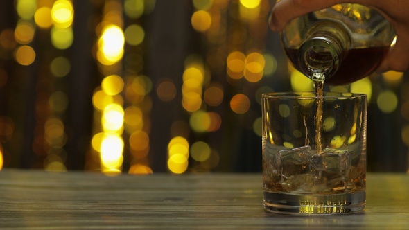Pouring of Golden Whiskey, Cognac or Brandy From Bottle Into Glass with Ice Cubes. Shiny Background alt