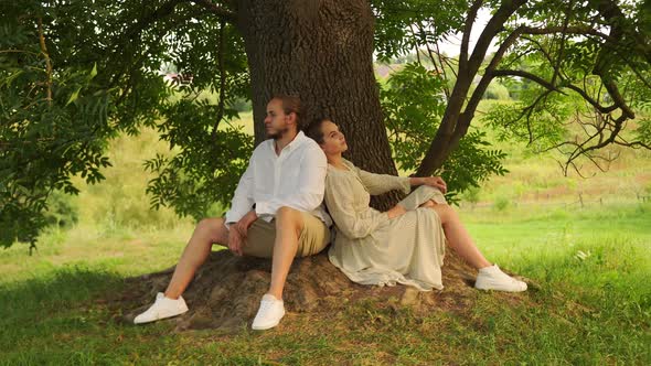 Young Couple Love Sitting with Their Backs to Each Other Under Tree in the Park