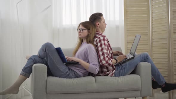 Side View Wide Shot of Young Man and Woman Sitting on Couch Leaned at Each Other and Using Laptops alt