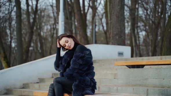 Luxury girl sitting on bench. Attractive young woman in fur coat enjoys fresh air in the park alt