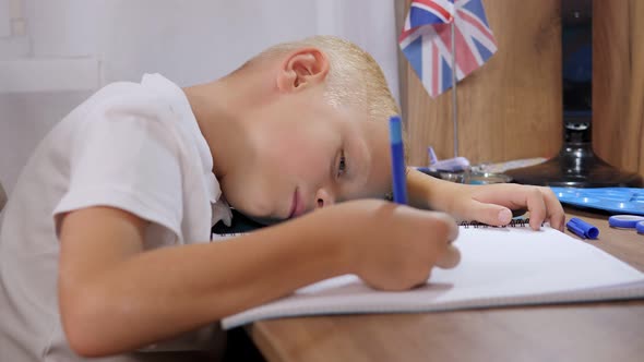 Tired Little Boy Sitting at the Table with His Head on Arm Writes in Notebook alt