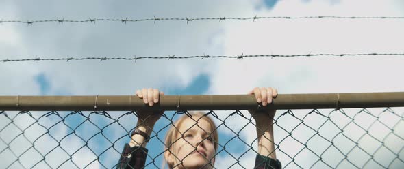 Young woman climbs metal fence and she is looking around over the fence alt