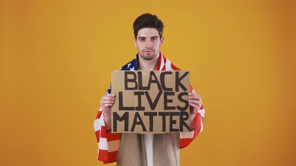 Fellow Wrapped in Flag of USA is Raising Up Cardboard Poster with Inscription Black Lives Matter alt