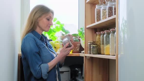 Storing Food in Pantry Middleaged Woman in Kitchen Near Wooden Shelf with Jars of Cereals alt