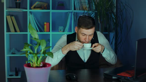Young Man Drinks Coffee Sitting in Armchair Before Computer in Office alt