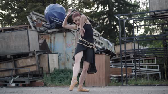 Wide Shot of Slim Elegant Ballerina in Black Dress Bound with Rope Dancing on Urban Slum alt