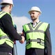 Positive Caucasian Men in Hard Hats and Uniform Shaking Hands at Background of Clear Sky Outdoors