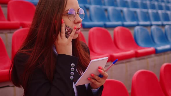 Young Woman in Glasses with Notepad Pen Talking on Mobile Phone Sitting on Stadium Bleachers alt