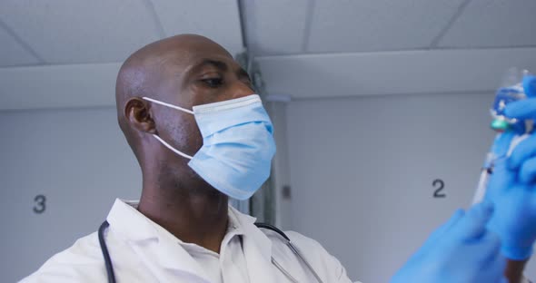 African american male doctor wearing face mask filling the syringe with covid-19 vaccine at hospital alt