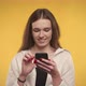 Young Adult Caucasian Woman is Typing on Her Smartphone on a Bright Yellow Background