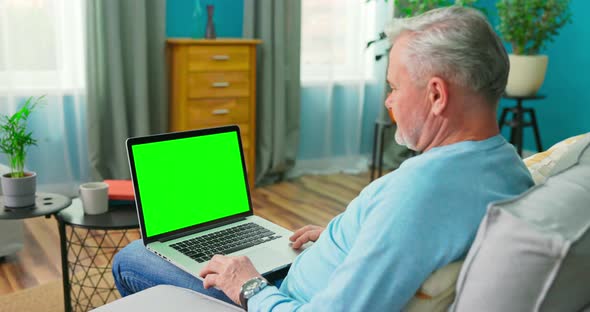 Older Man at Home Sitting on a Couch Works on a Laptop Computer with Green alt