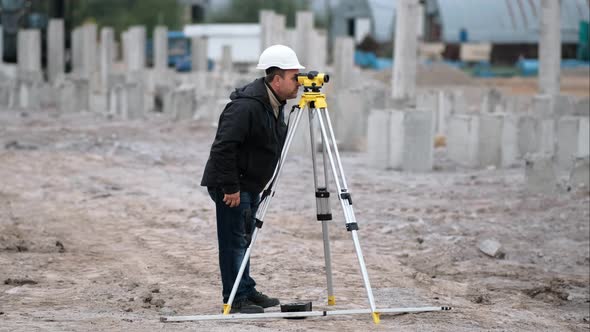 A gray-haired man in a white helmet looks into a yellow theodolite on a construction site