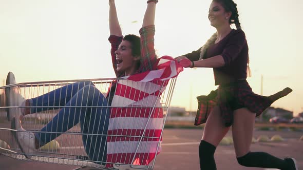 Young Hipster Teen Girls Having Fun at the Shopping Mall Parking Riding in Shopping Cart Holding the alt