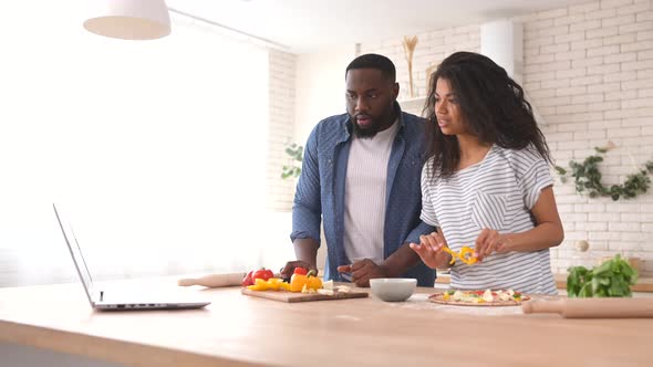 Happy Multiracial Couple Preparing Meal Together in Front of a Laptop alt