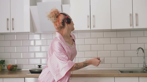 Happy Young Woman with Pink Hair Dancing in Kitchen Wearing Pink Pajamas and Listening to Music with alt