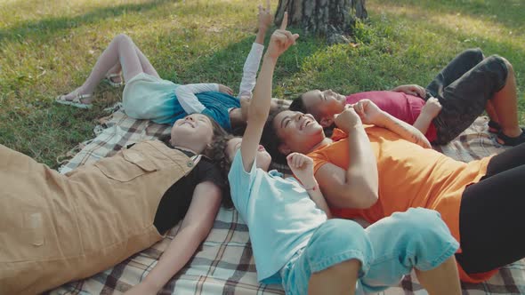 Children and Young Woman Lying Back Looking Up Clouds in Public Park alt