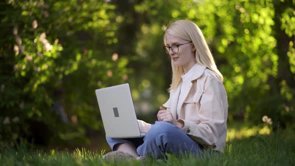 A Young Beautiful Woman in Light Clothes Sits on the Green Grass and Talks By Video Call Via a