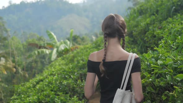 Young Woman with Bag Walks Along Path and Smells Tea Leaf alt