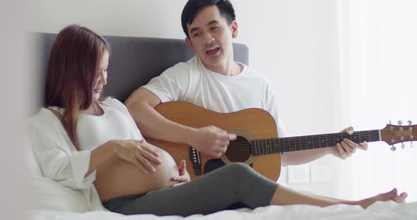 Asian husband and pregnant wife spend time together in the bedroom and husband play guitar.