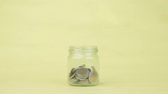 Woman hand putting the coin into a clear glass jar on yellow background, Saving money concept.