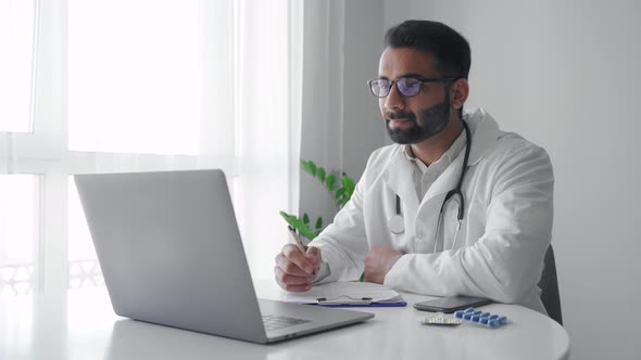 Indian Man Physician Wearing White Coat Video Calling Distant Patient on Laptop alt