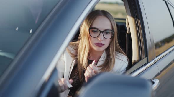 Business Lady Looking in Rear View Mirror and Applying Red Lipstick in Car alt